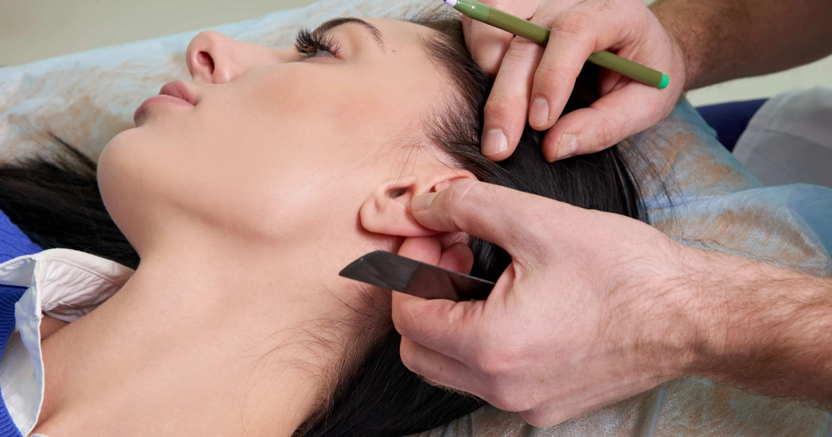 Close-up of a woman’s side profile as a professional examines her ear and jawline, marking for Otoplasty Surgery in Providence, UT.
